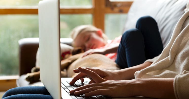 Woman uses a laptop while a child sleeps on a sofa nearby